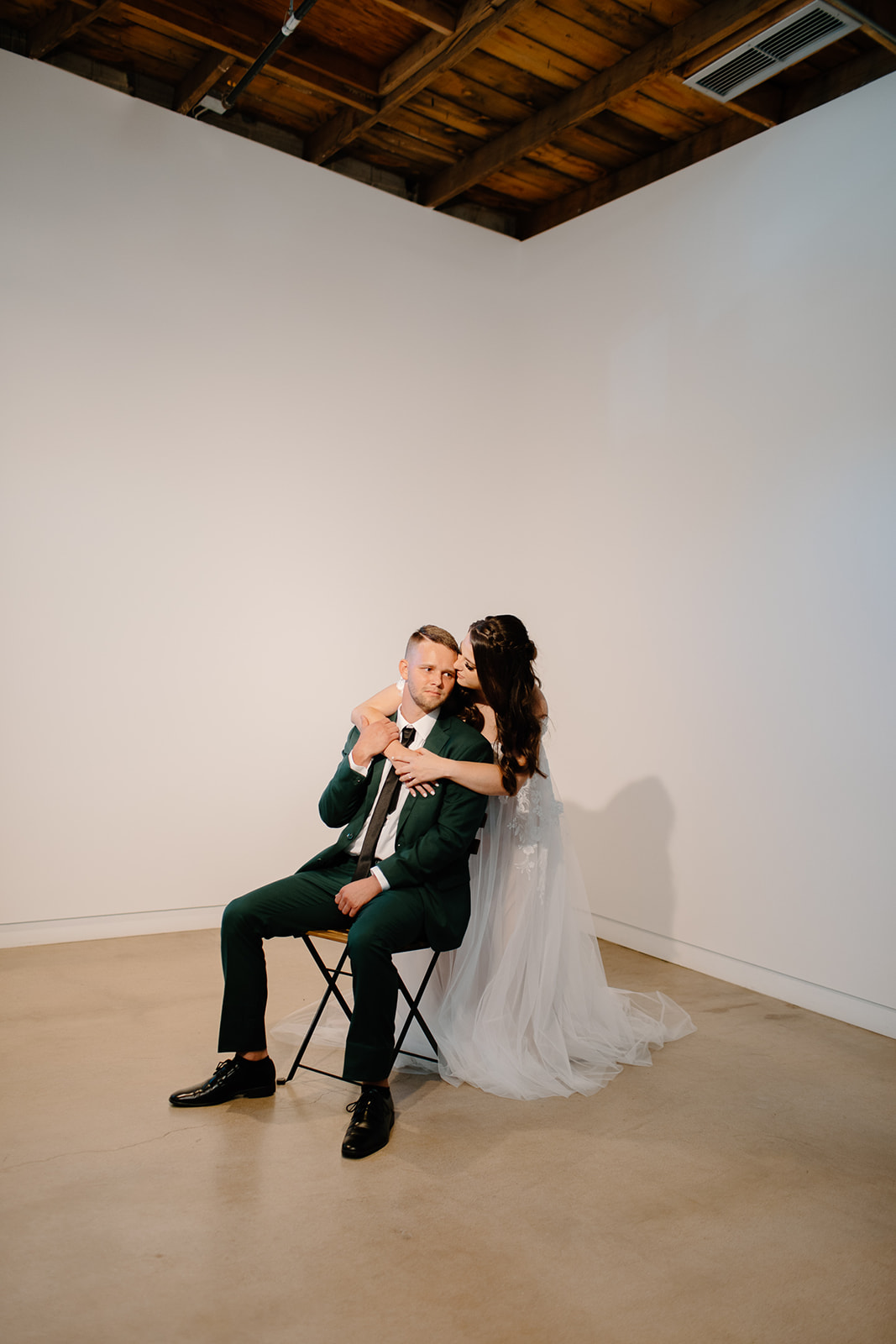 Groom sitting in chair, bride with arms over his shoulders giving him a kiss on the cheek.